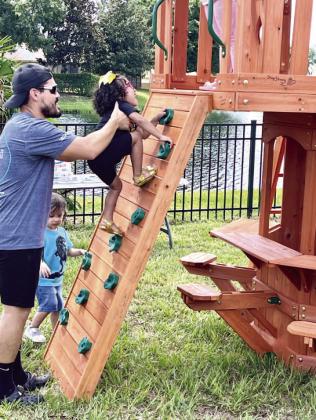 Volunteers from Roc Solid and Amazon built the playset in the Aguilars’ back yard. Once built, Denys Aguilar helps Devyn conquer her new rock-climbing wall. PHOTO/DEBBIE DANIEL
