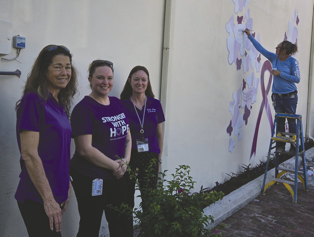 Help Now of Osceola County’s Tammy Douglass, Christina Montolvo and Andie Hinman planted a new bougainvillea tree while artist Jules Ross touches up a domestic violence awareness mural she first painted in 2021 in downtown Kissimmee in honor of Christina’s sister Nicole Montalvo, who was a high-profile violence victim in 2019. (Photos/Ken Jackson)
